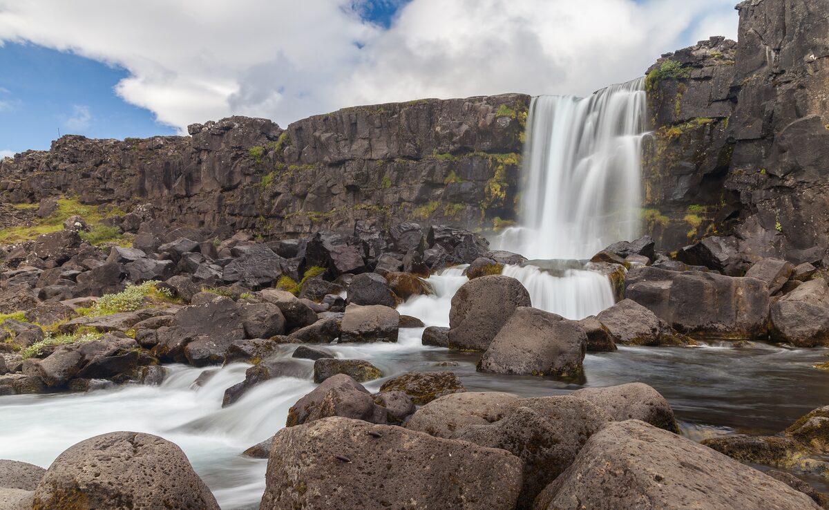 Öxarárfoss falling over a dark basalt cliff in Þingvellir National Park, with boulders in the foreground and blue sky above
