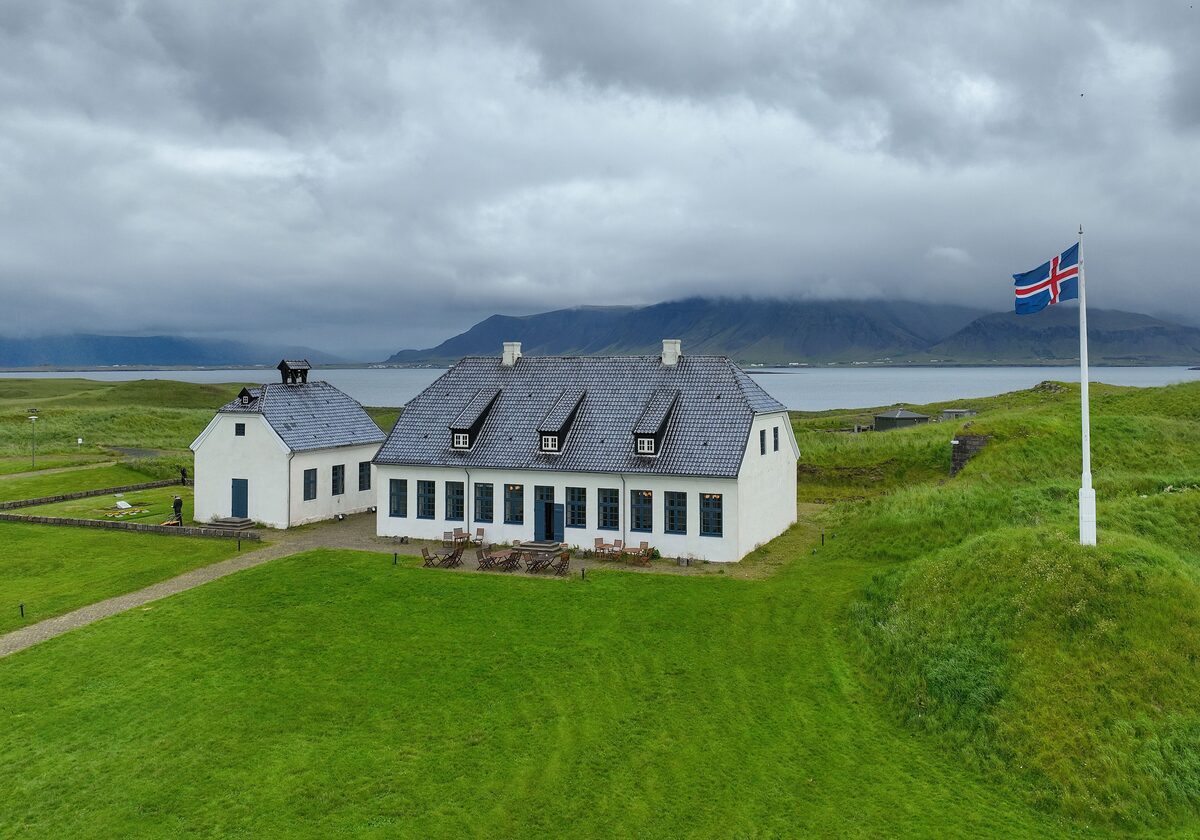 A white house with a dark roof and a flag on the grassy island of Viðey