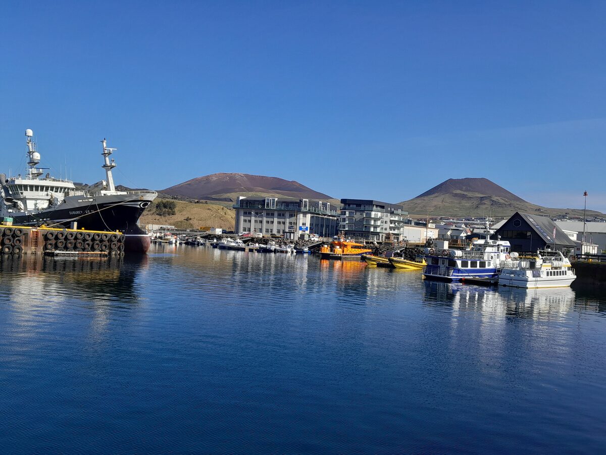 Heimaey harbour on a clear day, with fishing boats at the quay and the Eldfell and Helgafell volcano cones behind the town