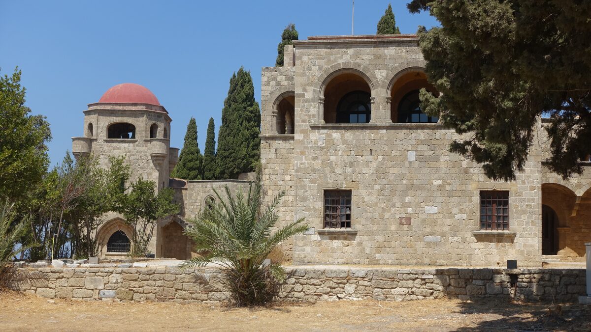 The stone buildings of Filerimos Monastery with a red-domed chapel tower, cypresses and a palm in the courtyard