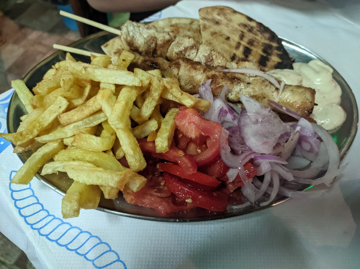 A pork souvlaki plate with grilled meat, fries, sliced tomato and onion, and tzatziki on the side
