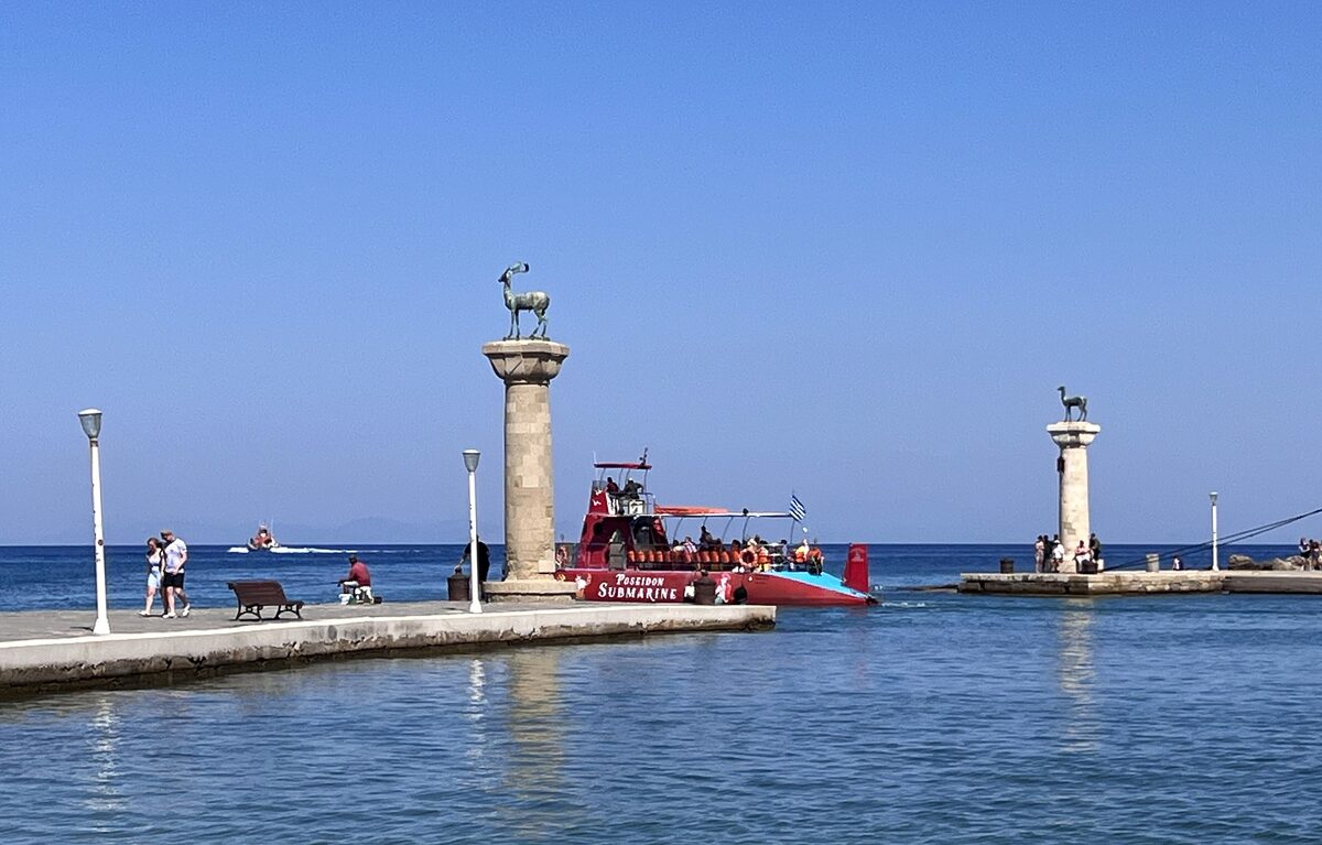 The two bronze deer on their columns at the entrance to Mandraki harbor, with a red excursion boat beyond