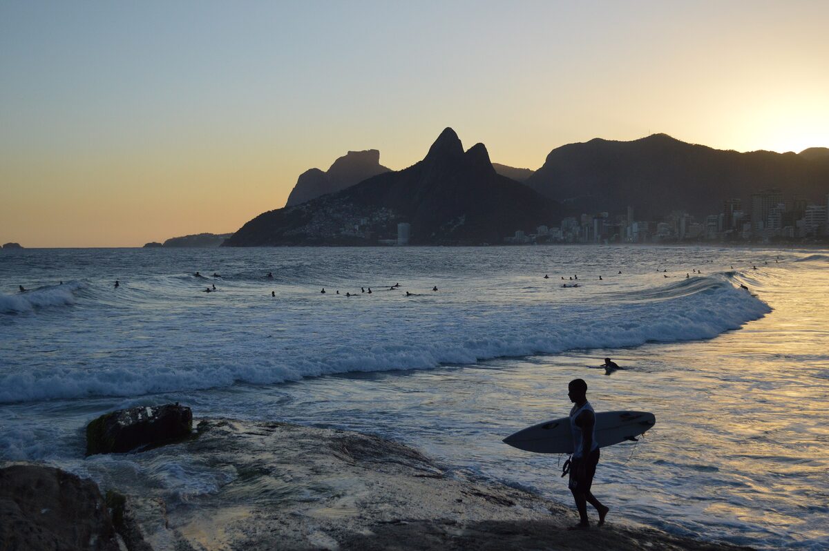 A surfer walking out of the surf at Arpoador at sunset, with the mountains in silhouette