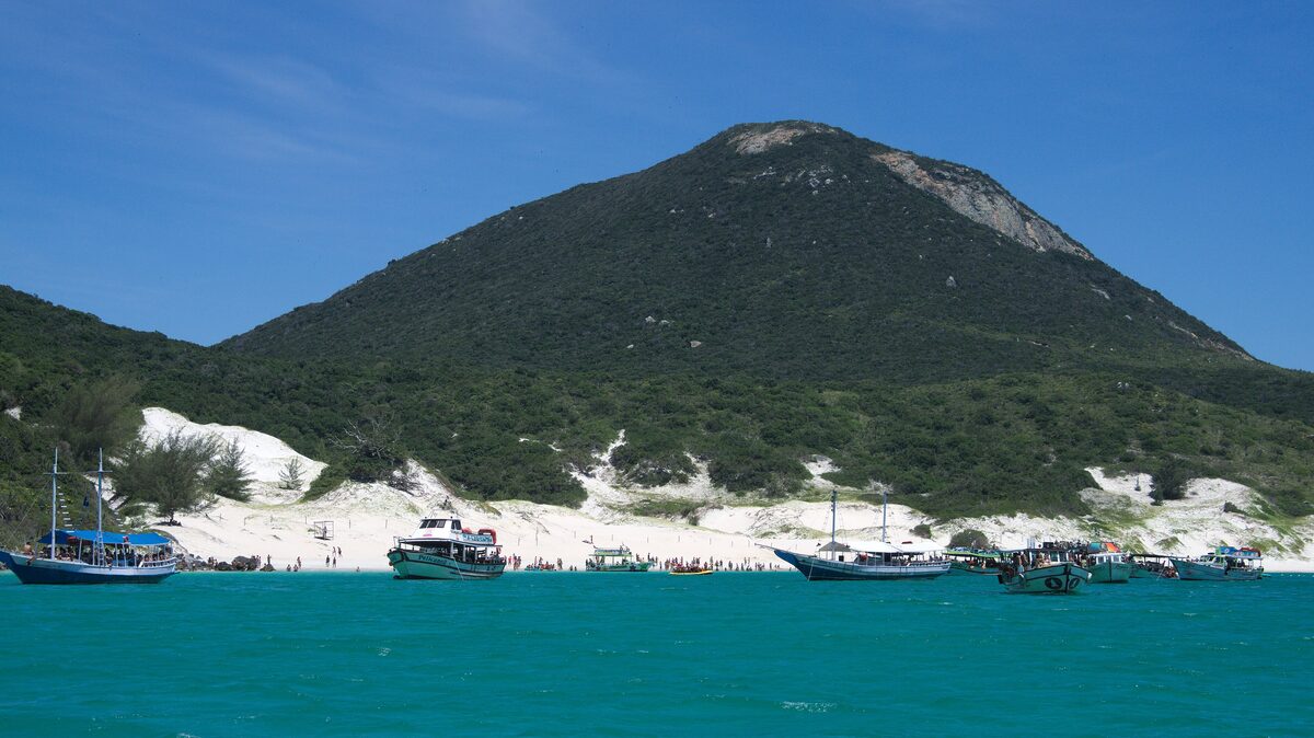 Boats anchored in turquoise water off a white-sand beach below a green hill on Ilha do Cabo Frio, Arraial do Cabo