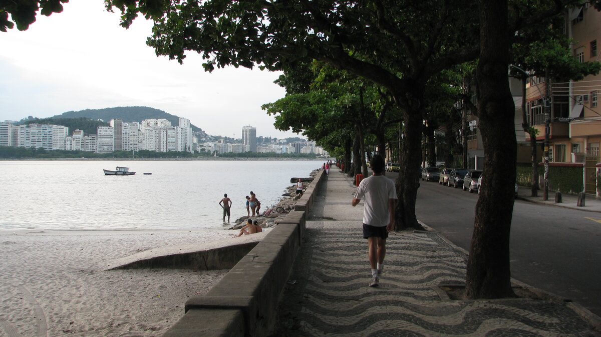 The Urca sea wall and its black-and-white wave pavement, with a walker seen from behind, the bay and the Botafogo skyline across the water