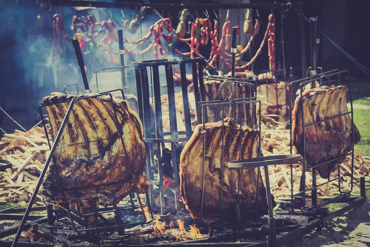 Large racks of ribs roasting upright on iron frames beside an open wood fire, with sausages hanging behind
