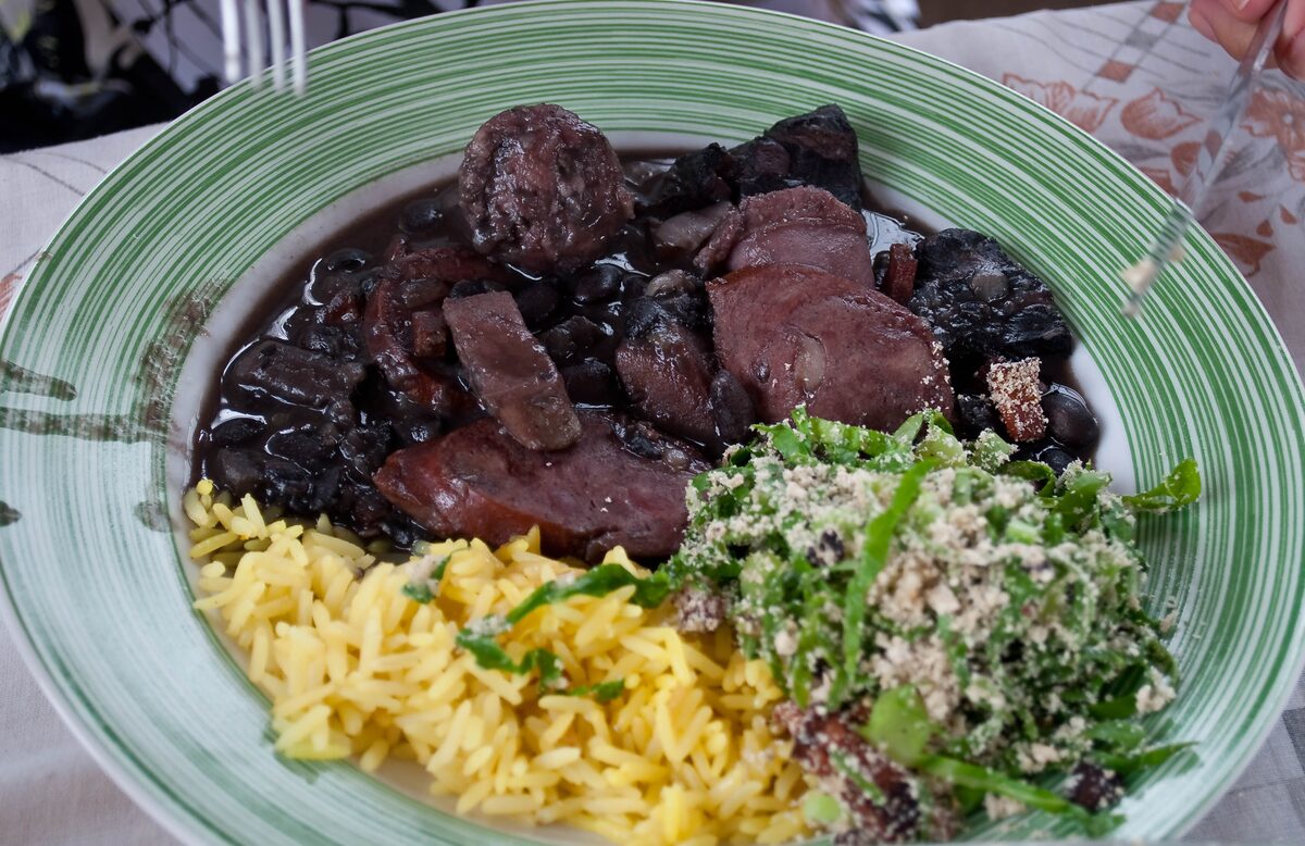 A plate of feijoada with black beans and pork, yellow rice, collard greens and farofa on a green-rimmed plate