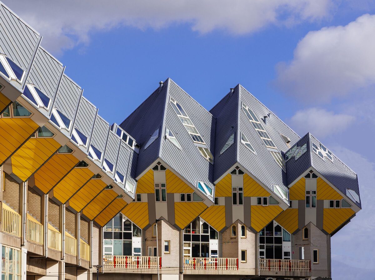 A row of yellow and gray Cube Houses tilted on their corners on top of concrete pillars, under a blue sky with clouds