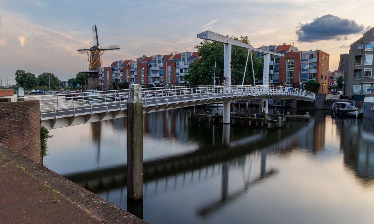 A white drawbridge over a still canal in Delfshaven at sunset, with a windmill and brick houses reflected in the water