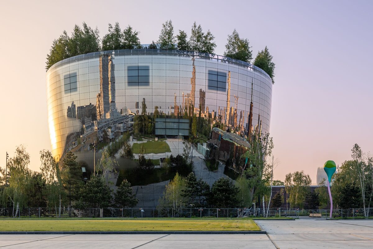The round, mirrored Depot Boijmans Van Beuningen with trees on its roof, reflecting the surrounding park and buildings in pink evening light