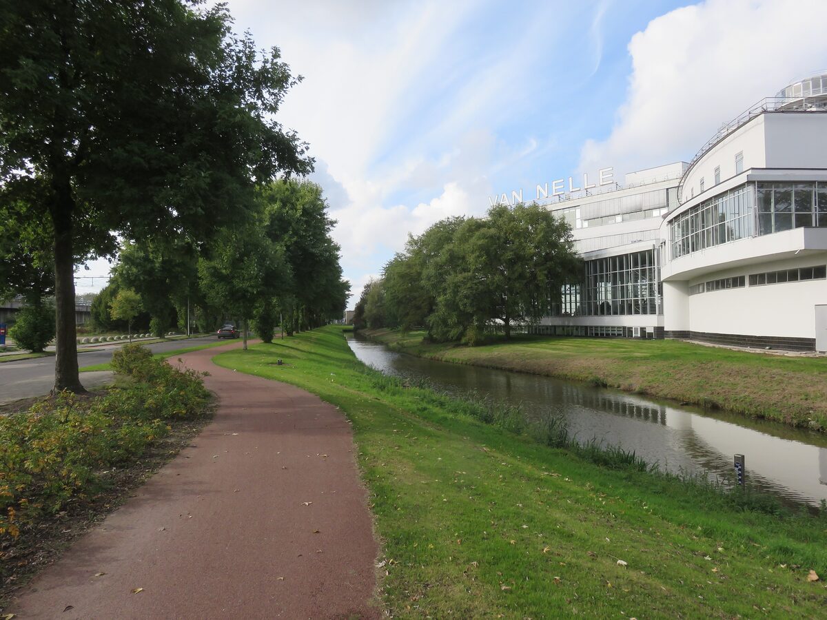 The long white glass and steel facade of the Van Nelle Factory beside a canal and a cycle path lined with trees