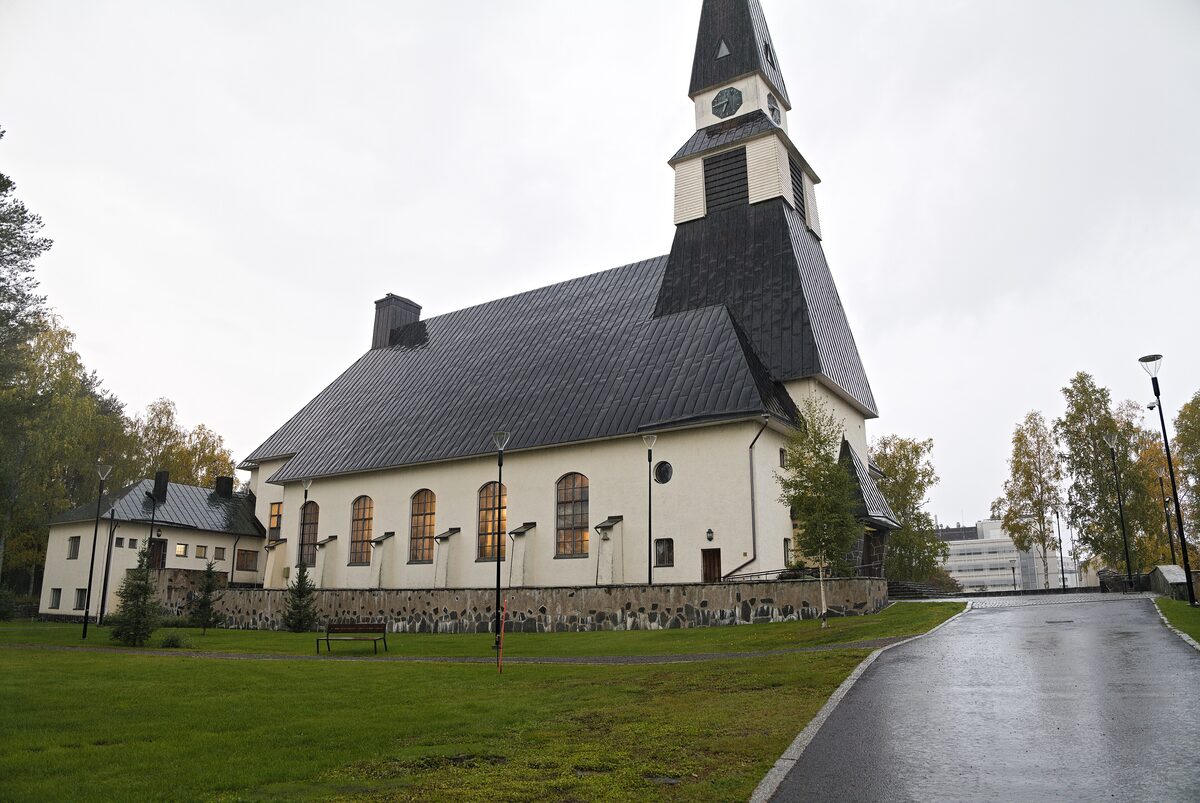 Rovaniemi Church, a pale church with a tall dark wooden spire, beside a wet road and green lawns