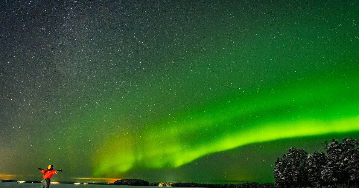 Green northern lights arching across a starry sky over a snowy, frozen landscape near Rovaniemi, with a person standing in the snow