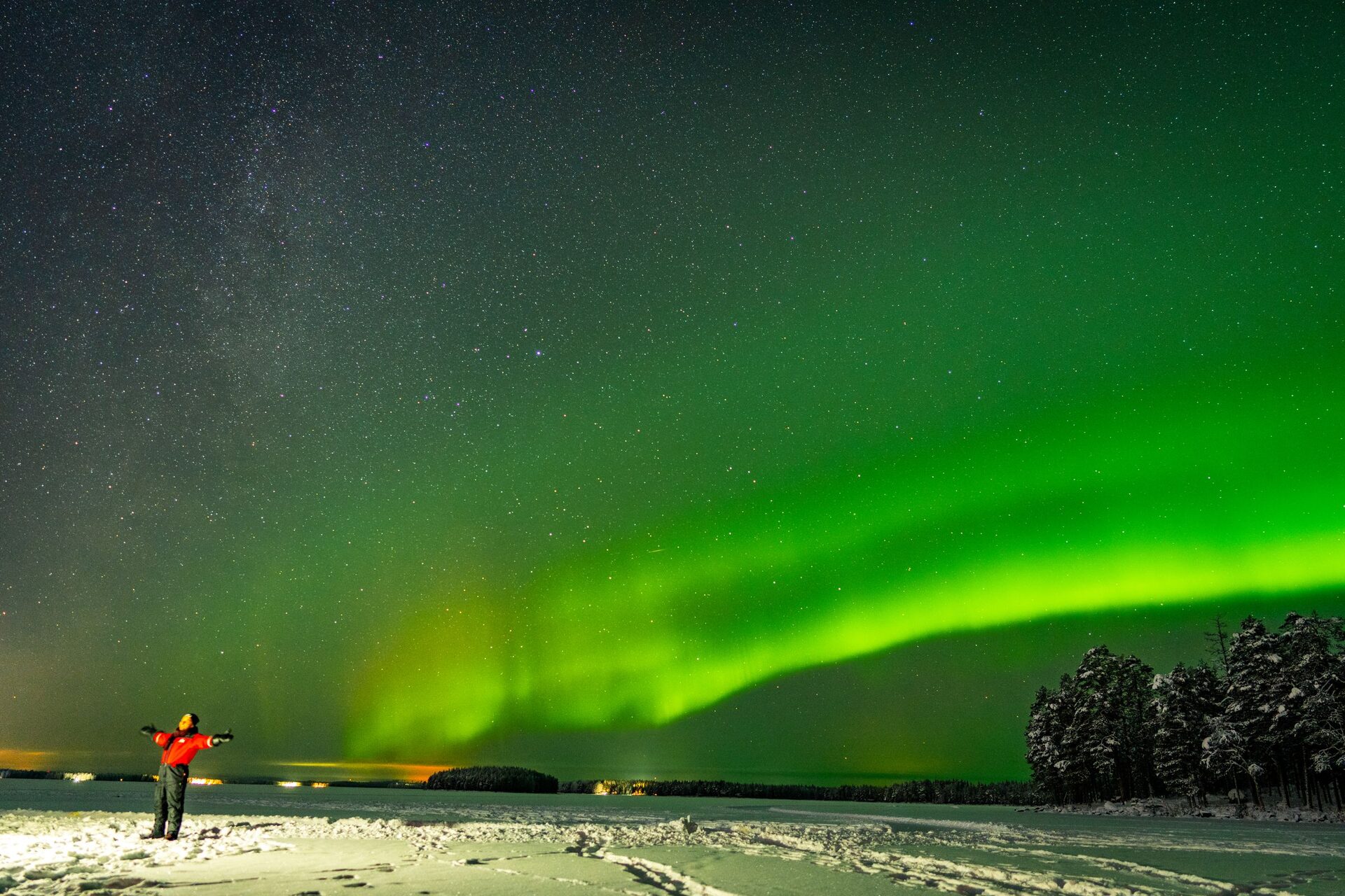 Green northern lights arching across a starry sky over a snowy, frozen landscape near Rovaniemi, with a person standing in the snow