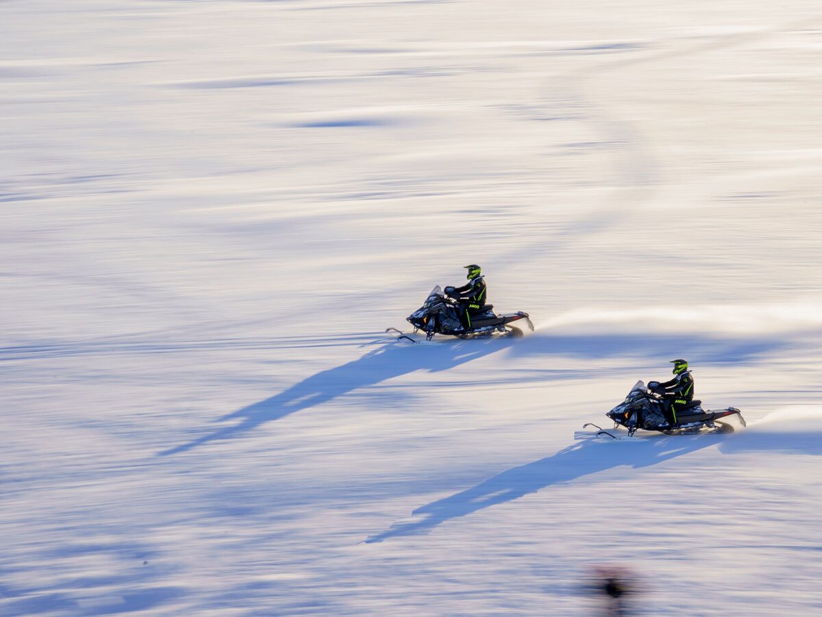 Two people riding snowmobiles fast across a vast, sunlit snowy field