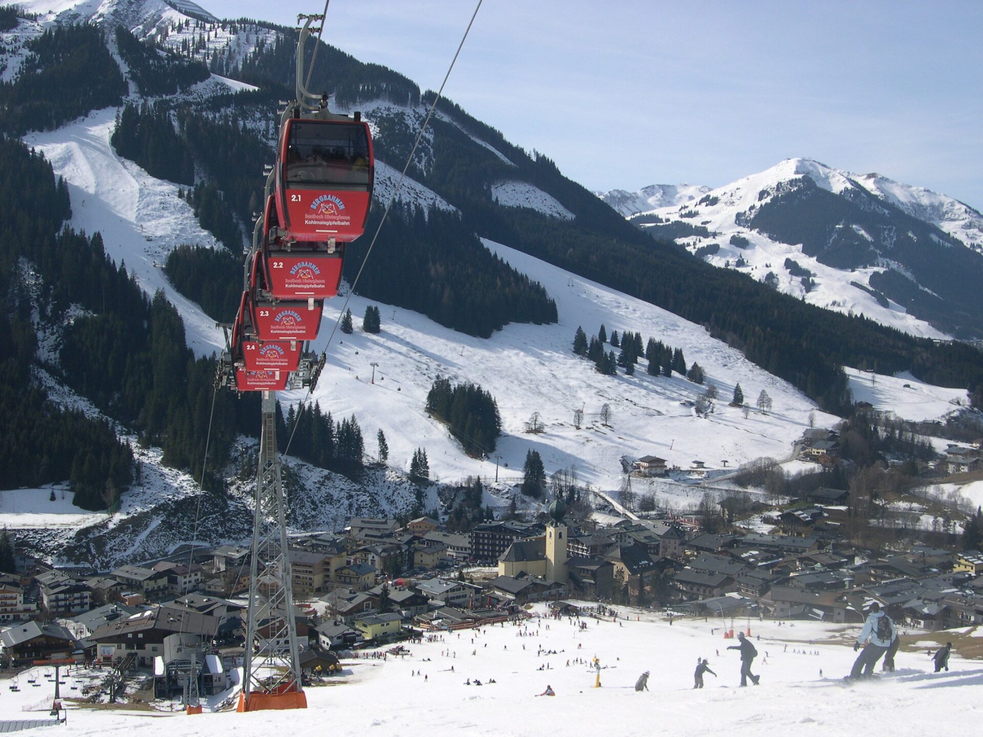 Red gondola cabins of the Kohlmaisbahn above Saalbach village, with the church and the snowy slopes in winter sun