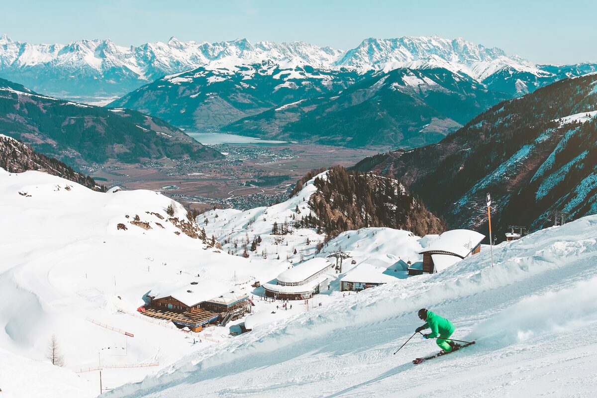 A skier in a green jacket carving down a snowy piste above Kaprun, with a mountain station and the Zell valley below