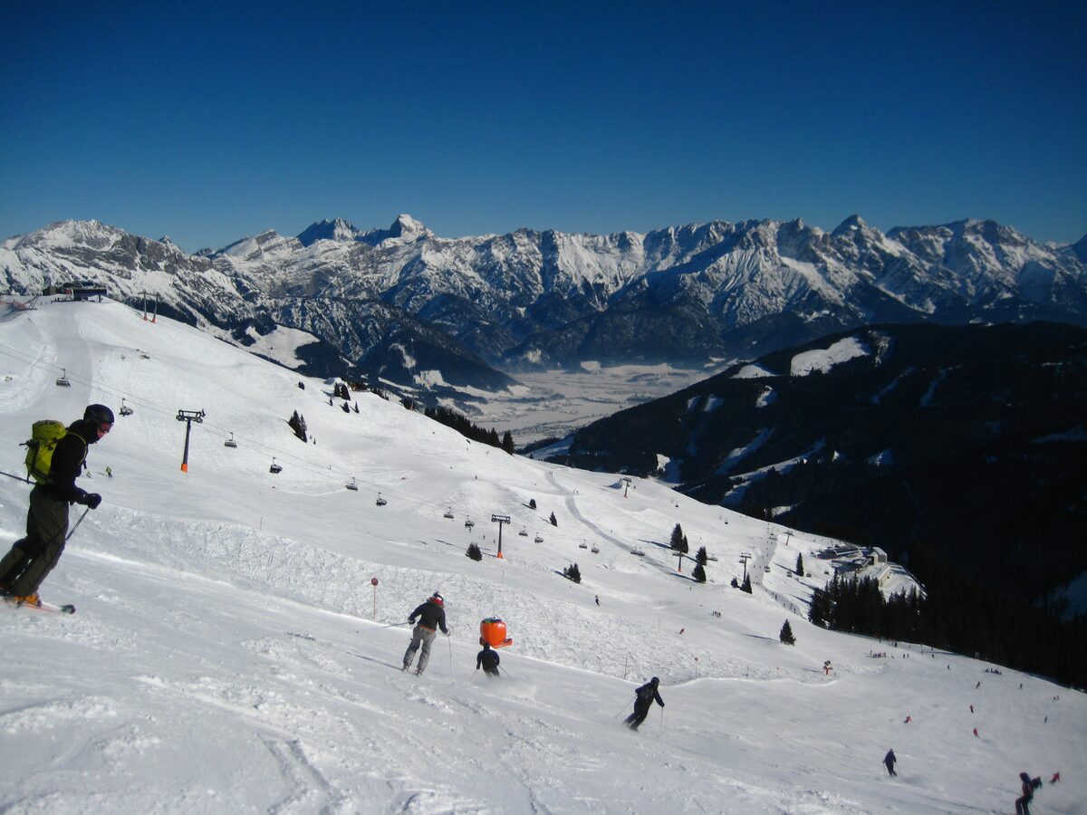 Skiers on a wide snowy piste at Leogang, with a valley and snow-capped peaks behind