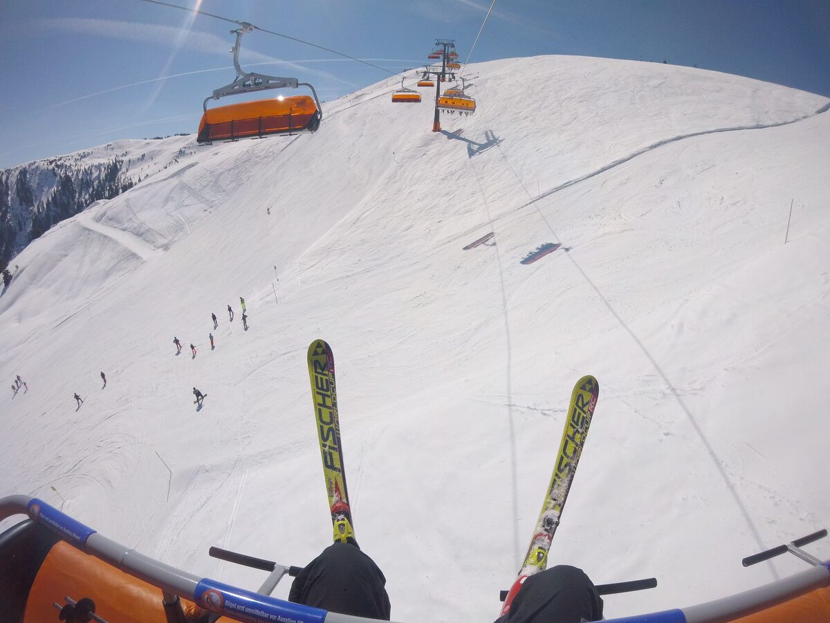 Skis hanging from a chairlift above a wide snowy slope in Leogang, with skiers far below and the next chairs ahead
