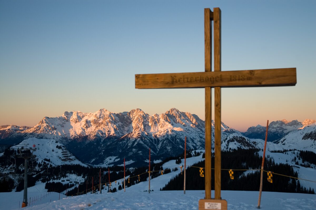 The wooden summit cross of the Reiterkogel at sunset, with the Loferer and Leoganger Steinberge glowing pink behind a snowy ridge