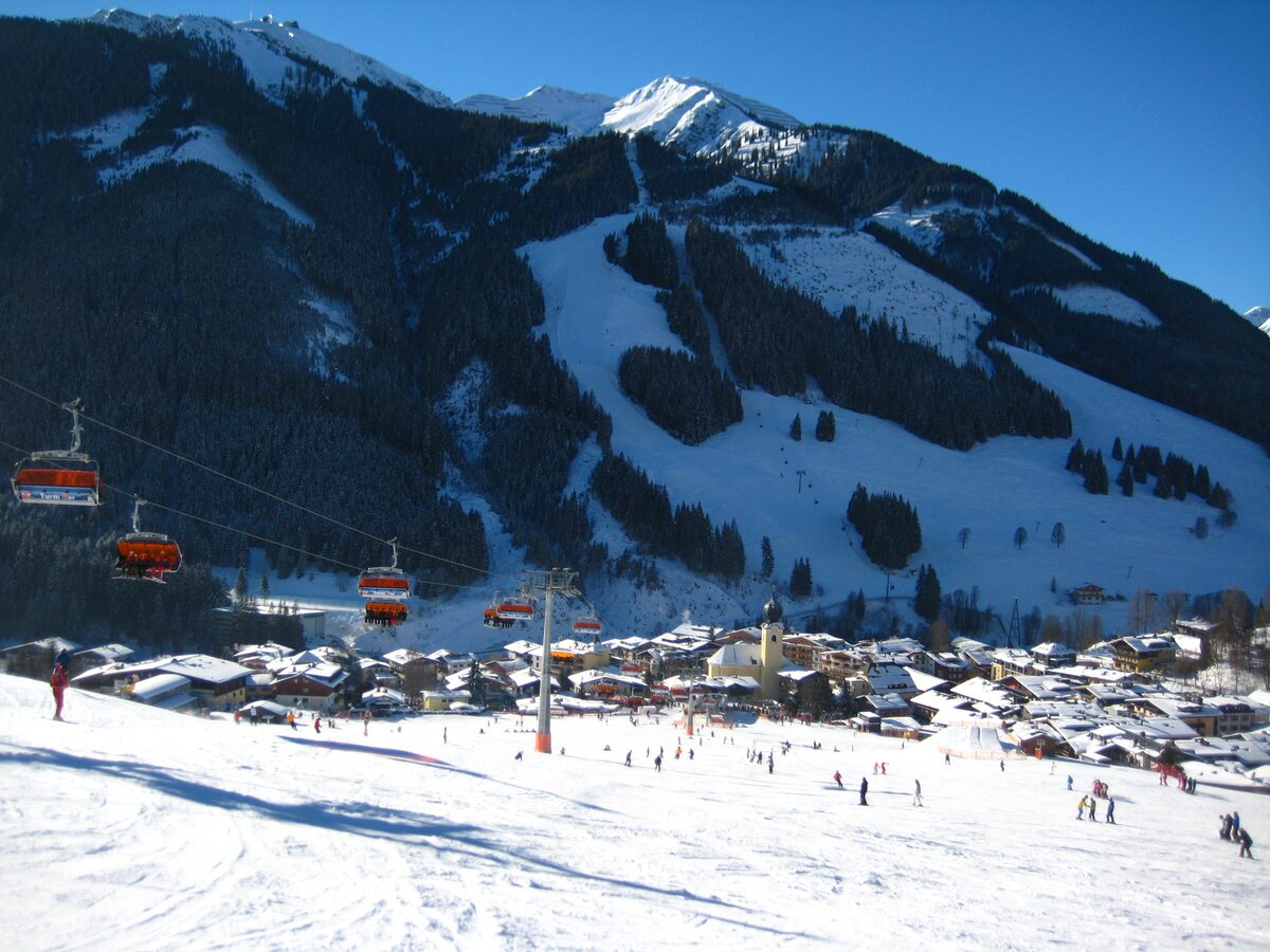 Saalbach in winter sun, with the church among the village roofs, skiers on the piste and red chairlift cabins climbing the slope above