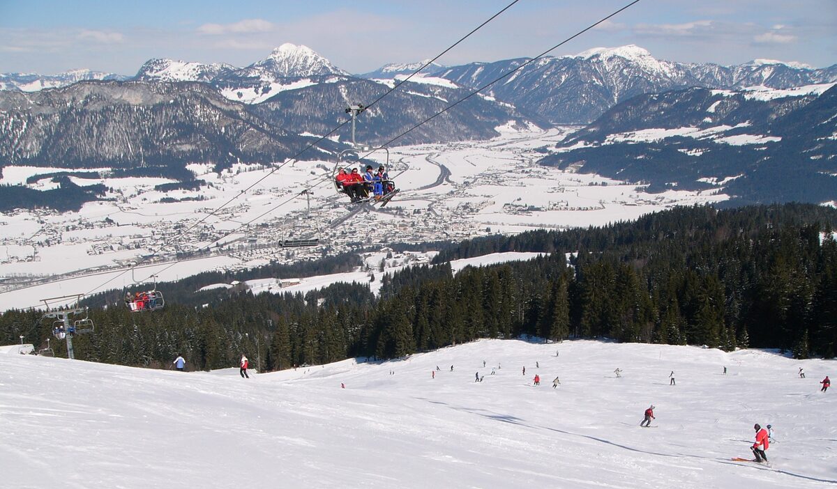 Skiers on a wide snowy slope in the Fieberbrunn ski area, with a chairlift overhead and a snowy valley and peaks behind