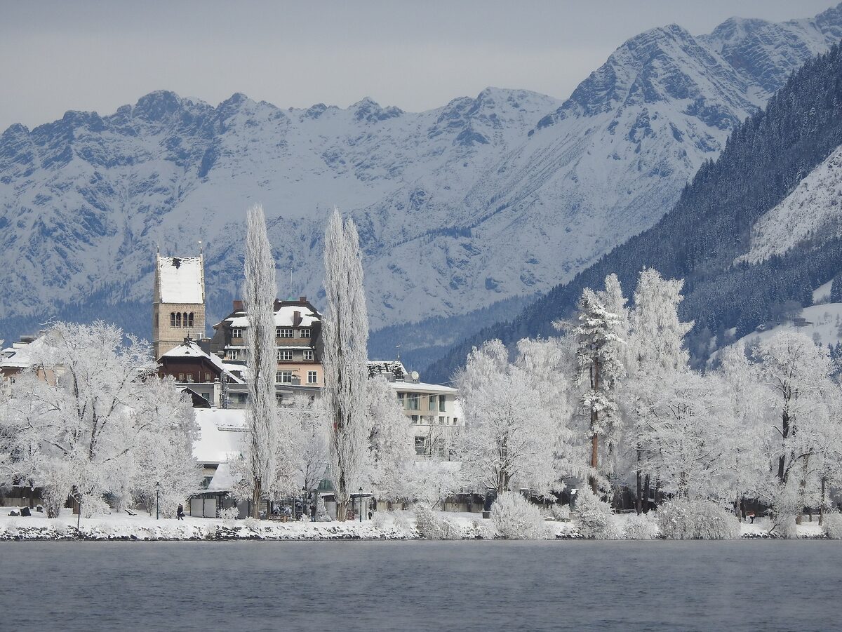 Frost-covered trees and a lakeside building on the Zeller See in winter, with snowy mountains behind