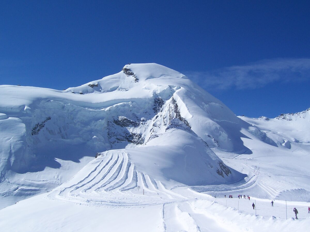 The Allalin glacier in winter, with groomed piste tracks and skiers on the snow below a white summit
