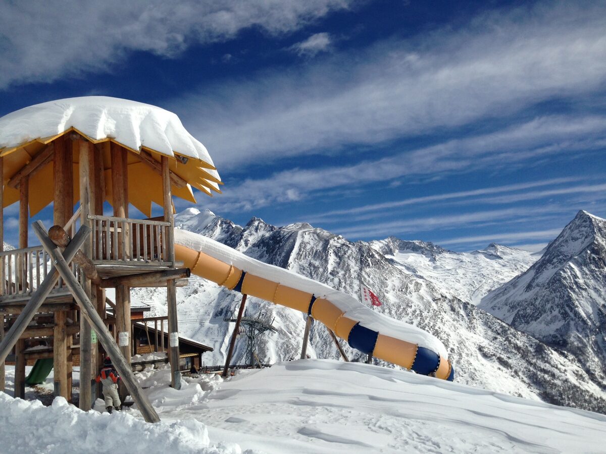 A wooden children's slide in deep snow at Kreuzboden, with snowy peaks behind