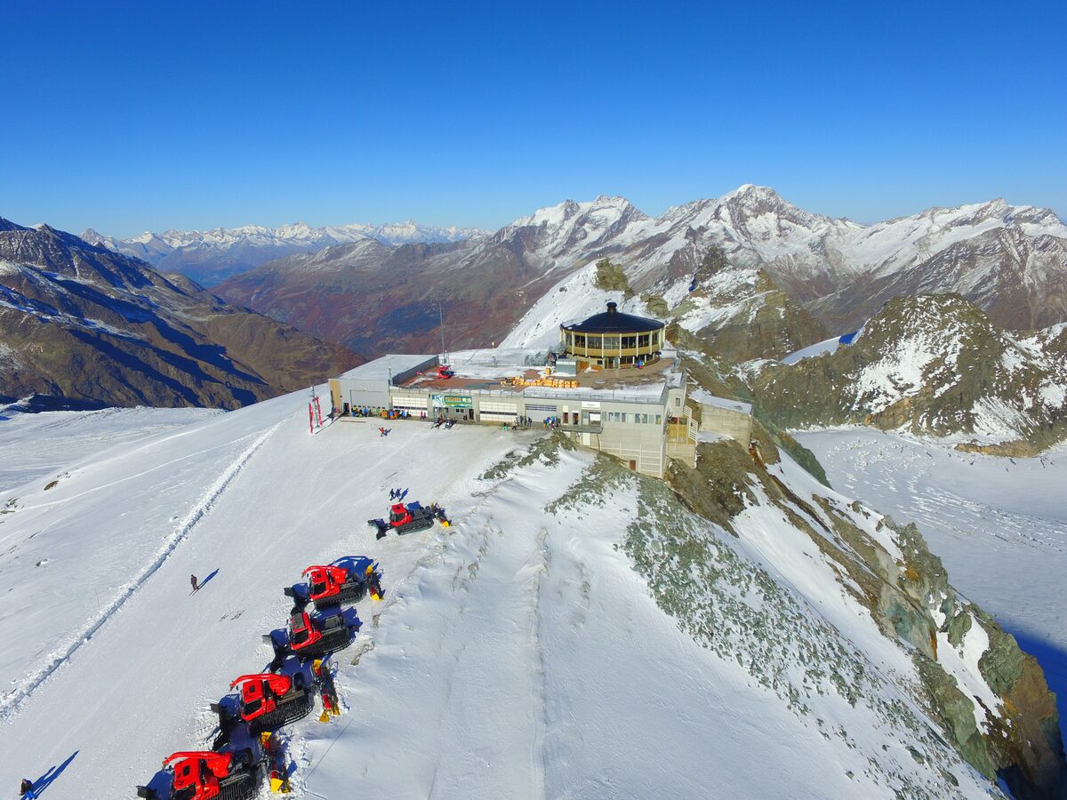 The round Allalin revolving restaurant on a snowy ridge, with groomers parked below