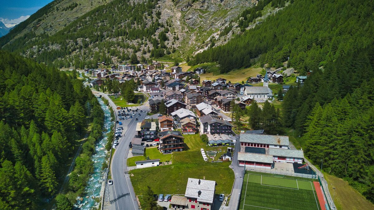 Aerial view of Saas-Almagell in summer, with the village houses beside the river and a sports field, under forested slopes