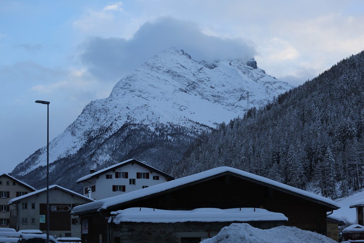 Snow-covered chalet roofs in Saas-Grund with the snowy Mittaghorn rising behind, its summit in cloud