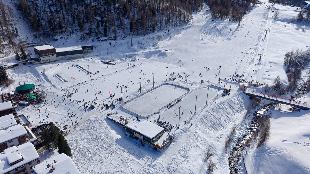 Aerial view of the snowy ski school area in Saas-Fee, with a skating rink and buildings among the pistes
