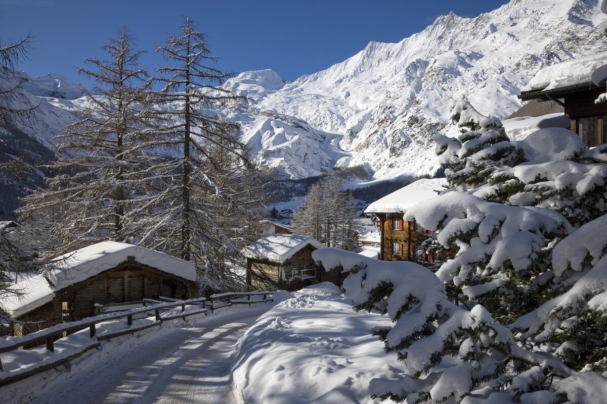 A snow-covered lane between wooden chalets in Saas-Fee, with pines and snowy peaks behind