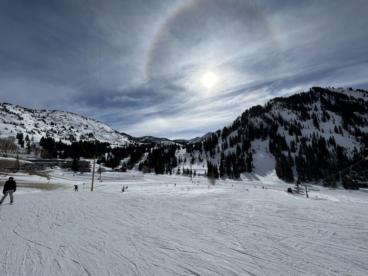 A groomed beginner slope at Alta under a pale winter sun with a halo, a lone skier at the left, a lift on the right and snowy ridges behind