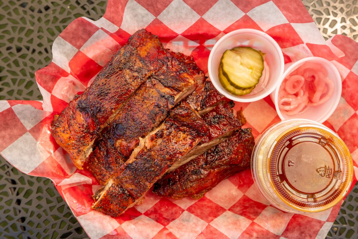 A rack of glazed barbecue ribs in a checkered paper basket with pickles and sauce