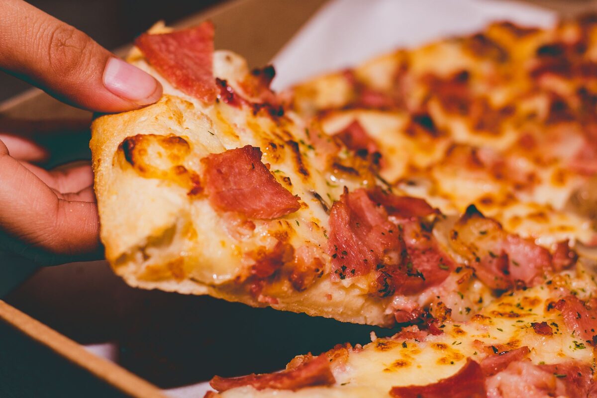 A hand lifting a slice of cheesy pizza with cured-meat topping from the box