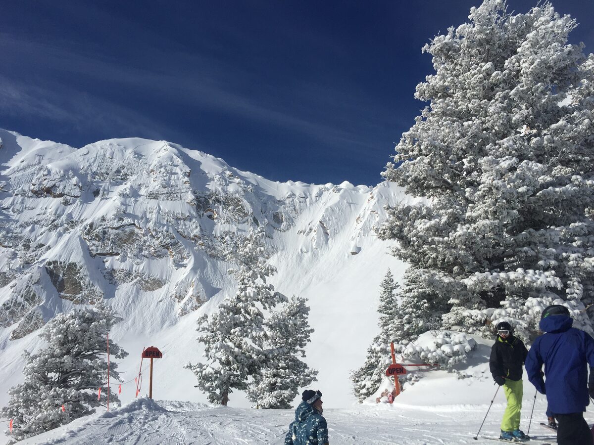Skiers pausing at the top of a steep, snow-plastered mountain face at Snowbasin, with frosted conifers and open-run signs