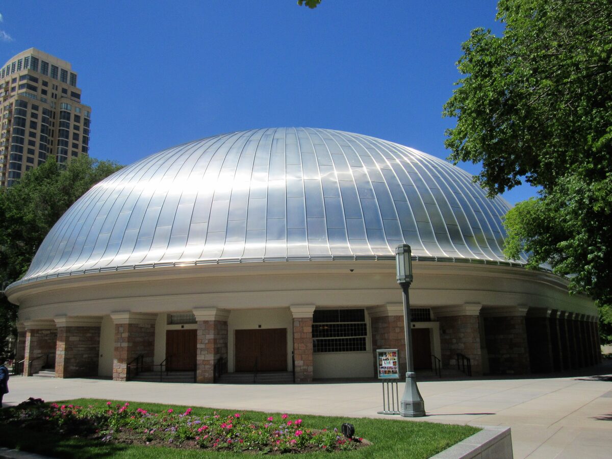 The silver-ribbed dome of the Salt Lake Tabernacle on Temple Square on a sunny day, with a flower bed in front and a downtown tower behind