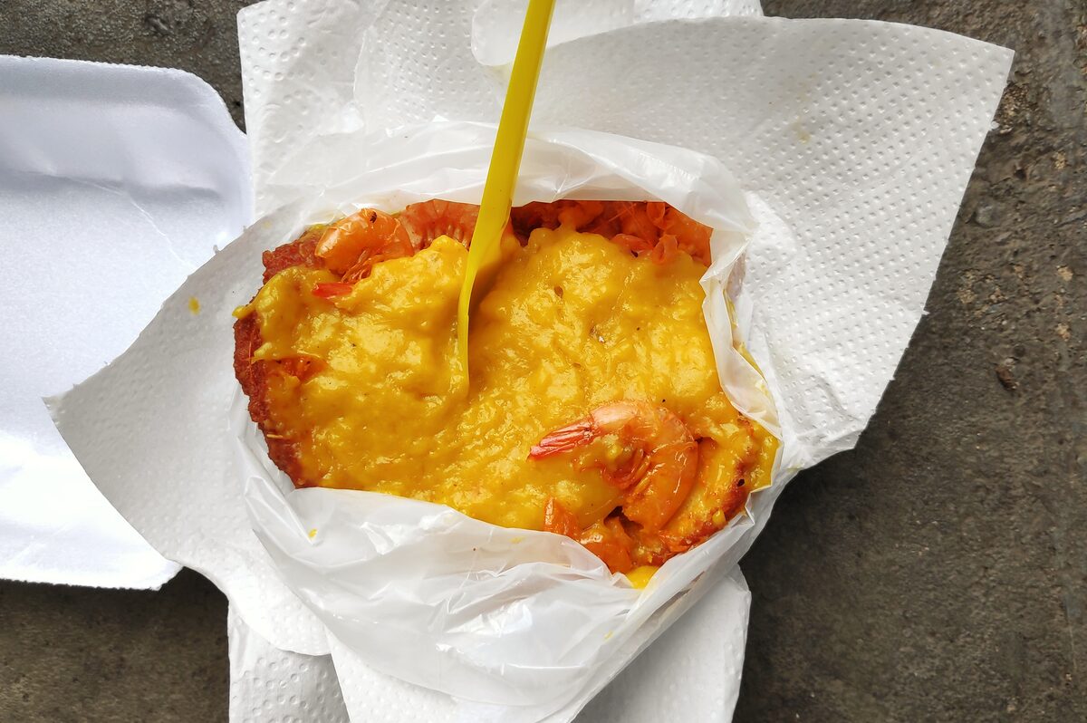 A takeaway acarajé split open and filled with yellow vatapá and a shrimp, on white paper