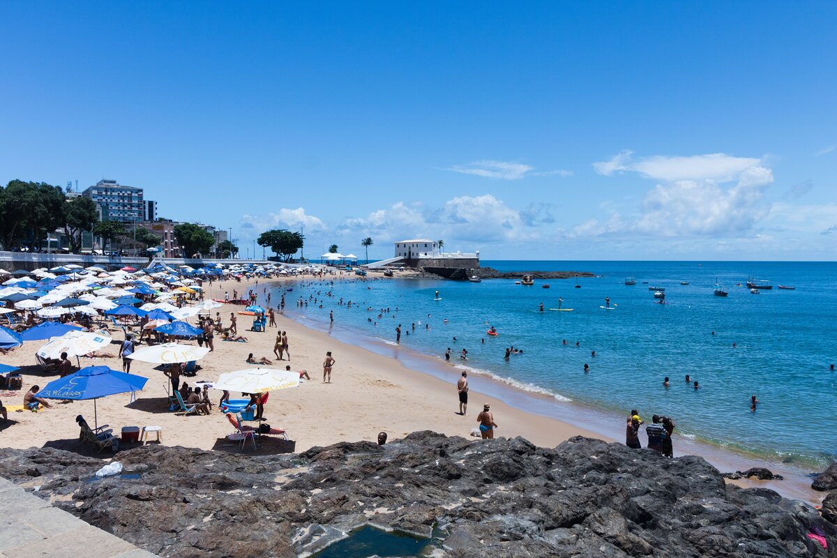 A busy city beach in daylight with blue umbrellas, swimmers and turquoise water, a fort on the point in the distance
