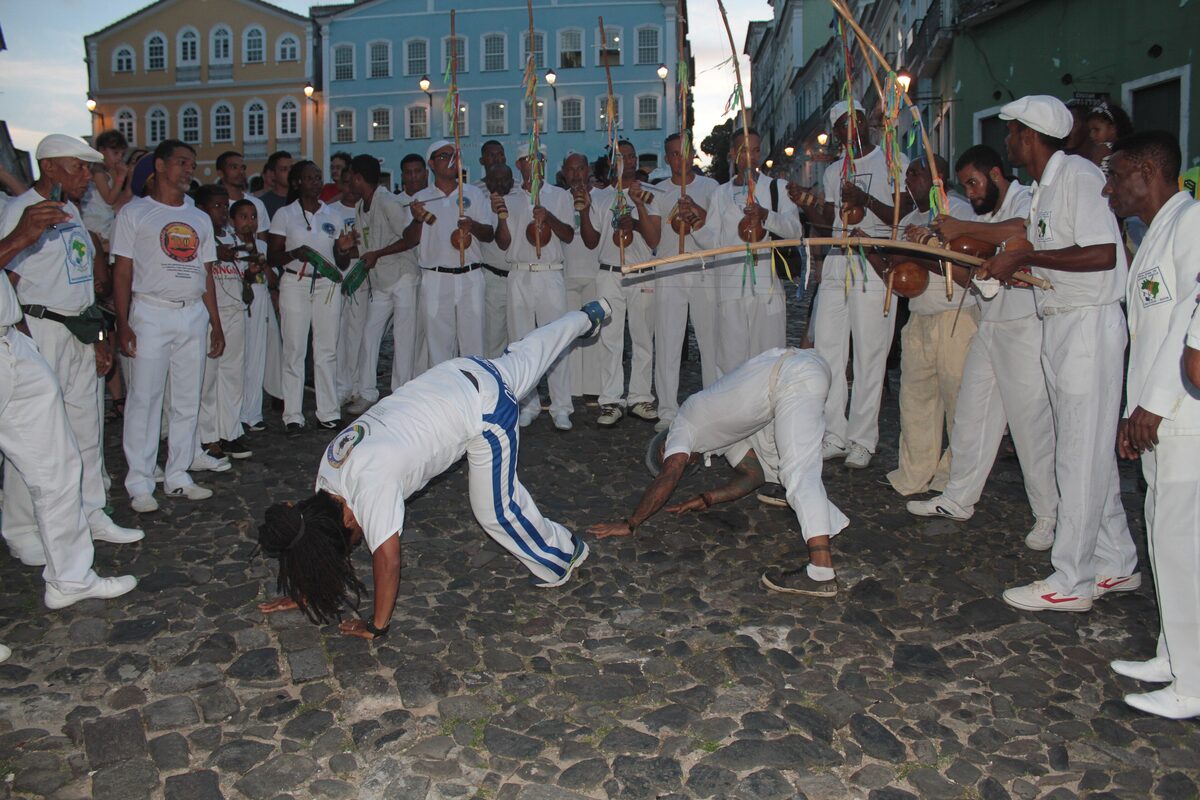 A capoeira roda at dusk on the cobblestones of the historic center, players in white around two capoeiristas, berimbaus raised