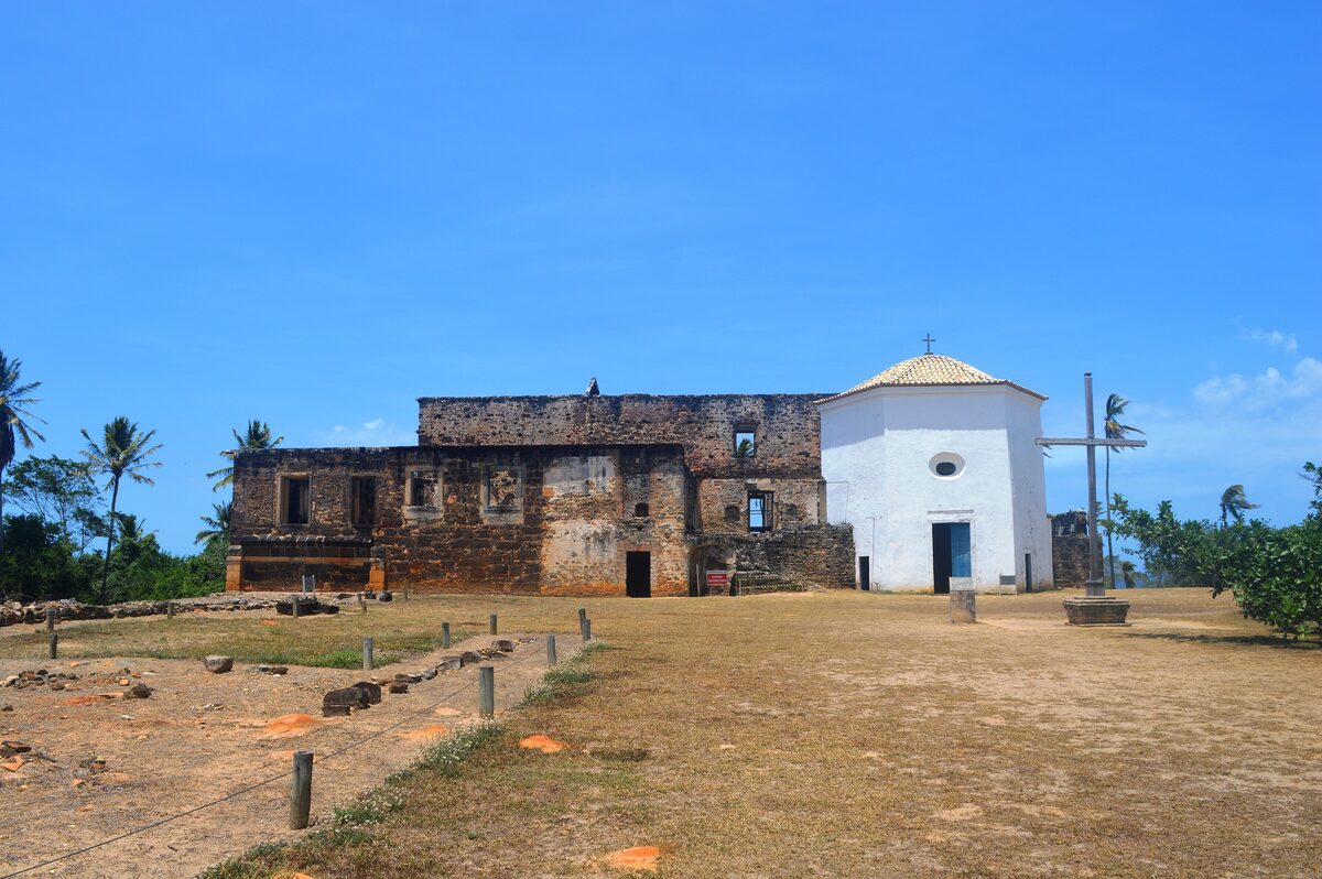 Ruins of the Garcia d'Ávila castle beside a small white chapel on open grass, with palm trees behind