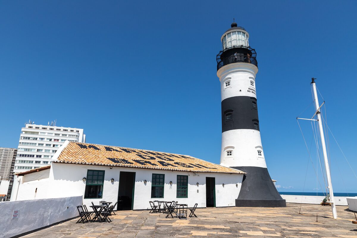 The black-and-white Farol da Barra lighthouse beside the white fort buildings, under a blue sky