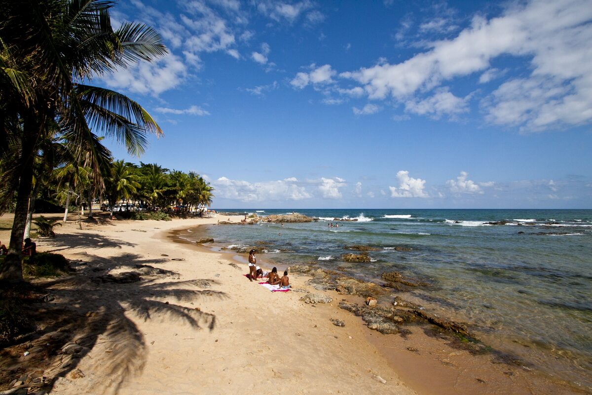 A sandy Itapuã beach framed by coconut palms, with rocks and blue sea under a partly cloudy sky