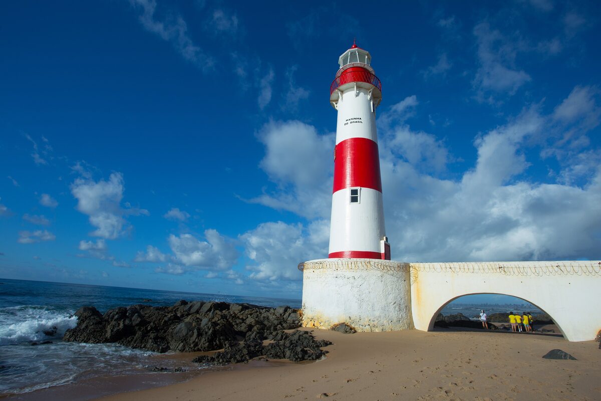 The red-and-white Itapuã lighthouse with a stone arch at its base, above dark rocks and sand