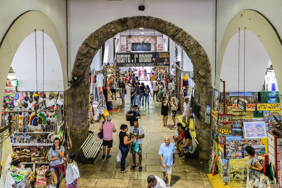 Inside the Mercado Modelo: shoppers walking between stalls of masks, paintings and souvenirs under a stone arch