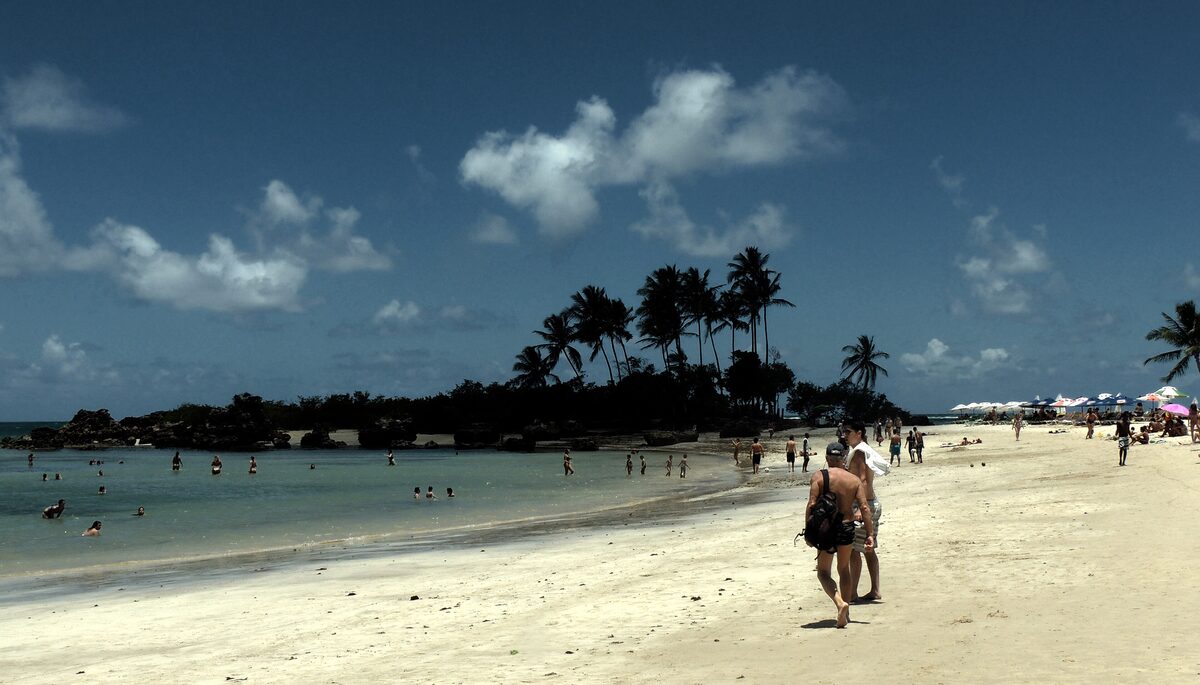 A palm-lined beach on Morro de São Paulo under a dark blue sky, with a rocky point and people walking on the sand