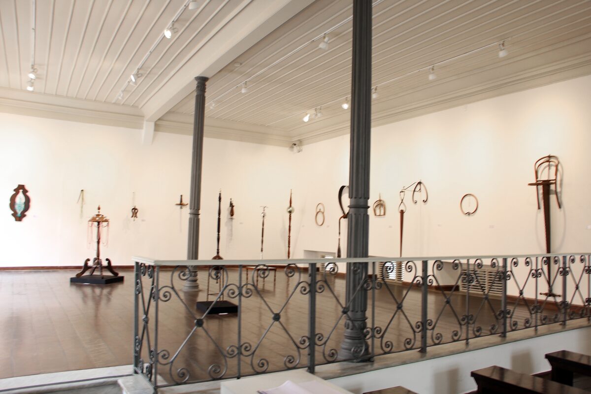A gallery room of the Afro-Brazilian Museum with metal objects on white walls and a wrought-iron railing