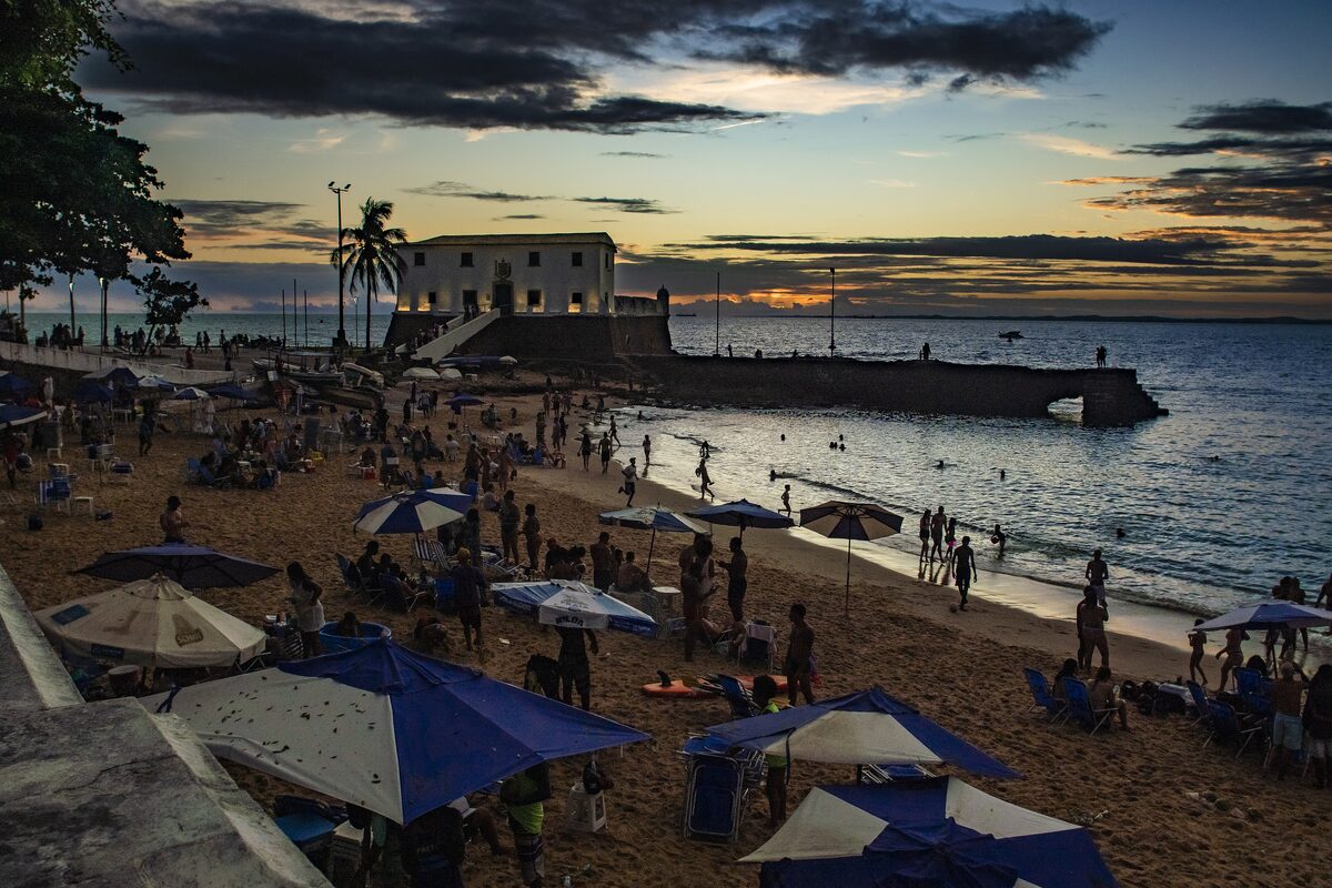 Porto da Barra beach at dusk: bathers and blue umbrellas on the sand, with the white Forte de Santa Maria against an orange-lit sky